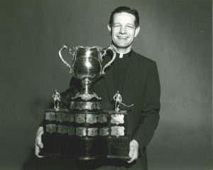Black and white photo. Fr. Bauer smiles and holds a trophy.
